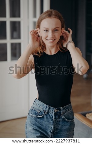 Smiling beautiful caucasian young woman fixing hairs at home, posing for photo portrait. Pretty female fashion model in casual clothes looking at camera with toothy smile, adjusting hair indoors.