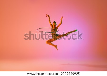 Young flexible teen girl rhythmic gymnast jumping isolated over pink background in neon light. Sport, beauty, competition, flexibility, active lifestyle. Individual performance