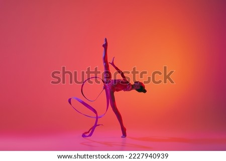 Exercise with ribbon. Young flexible teen girl rhythmic gymnast in motion, action isolated over pink background in neon light. Sport, beauty, competition, flexibility, active lifestyle. Performance