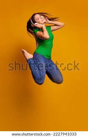 Jumping high, flying. Caucasian young woman's portrait on yellow studio background. Beautiful female model with headphones. Concept of human emotions, music, facial expression, youth, sales, ad.