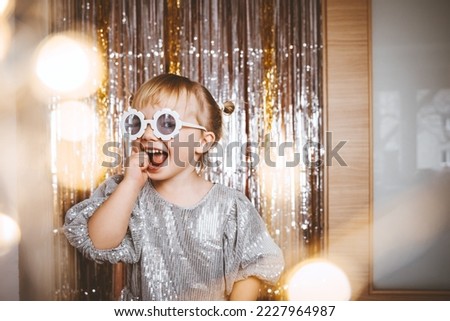 Happy little stylish girl in shiny dress having fun. Festive background with foil curtain decorations for kids birthday or fancy dress party, disco music or New Year. Celebration and Holiday concept. Royalty-Free Stock Photo #2227964987