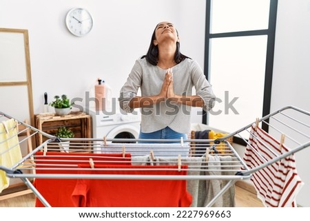 Young hispanic woman putting fresh laundry on clothesline begging and praying with hands together with hope expression on face very emotional and worried. begging. 