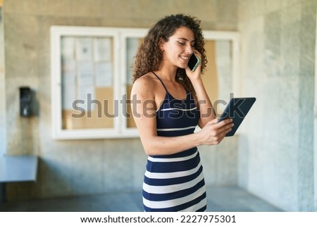 Young beautiful hispanic woman talking on smartphone using touchpad at street