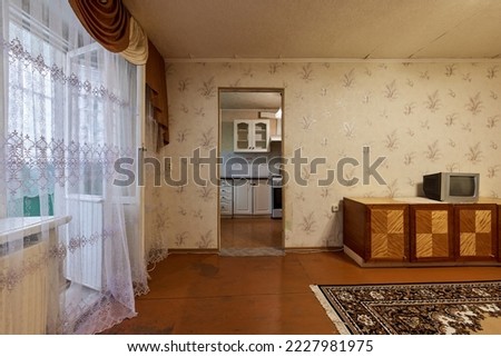 Example of Old Soviet Russian poor interior in Khruschev House. Aged sideboard, tv set, curtains, kitchen furniture. Shabby floor. Tattered wallpaper on wall. Carpet as decor. Apartment of pensioners. Royalty-Free Stock Photo #2227981975