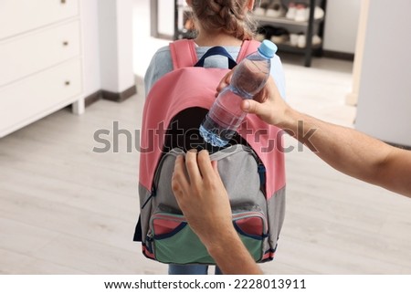 Father putting bottle of water into daughter`s backpack at home, closeup. Preparing to school Royalty-Free Stock Photo #2228013911