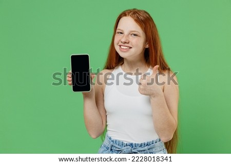 Little redhead kid girl 12-13 years old in white tank shirt hold use mobile cell phone with blank screen workspace area show thumb up isolated on plain green background. Childhood lifestyle concept.