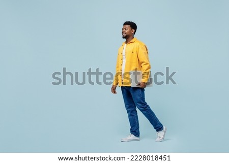 Full body young man of African American ethnicity 20s wearing yellow shirt hold use work on laptop pc computer isolated on plain pastel light blue background studio portrait. People lifestyle concept