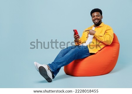 Full body young man of African American ethnicity 20s in yellow shirt sit in bag chair hold in hand use point finger on mobile cell phone isolated on plain pastel light blue background studio portrait Royalty-Free Stock Photo #2228018455