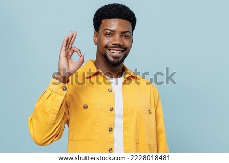 Young satisfied cheerful happy man of African American ethnicity 20s wear yellow shirt showing okay ok gesture isolated on plain pastel light blue background studio portrait. People lifestyle concept