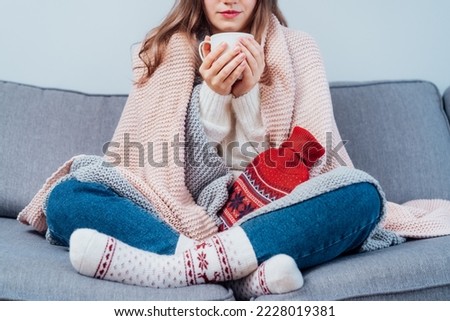 Woman freezes in wintertime. Young girl wearing warm woolen socks and wrapped into two blankets, holding a cup of hot drink and heating pad while sitting on sofa at home. Keep warm. Selective focus Royalty-Free Stock Photo #2228019381