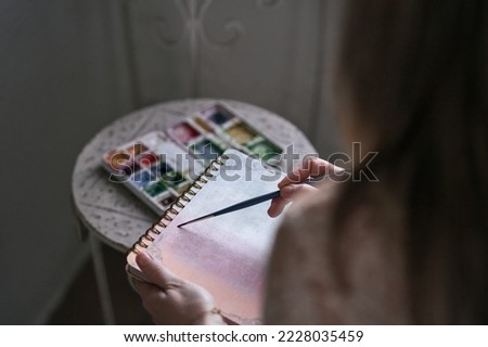 closeup of a caucasian mature woman's left hand holding a notebook while painting on it with a fine brush Royalty-Free Stock Photo #2228035459