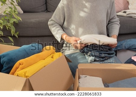 Woman sorting clothes and packing into cardboard box. Donations for charity, help low income families, declutter home, sell online, moving moving into new home, recycling, sustainable living concept Royalty-Free Stock Photo #2228061833