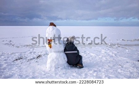 Seven years boy in knitted winter hat sits alone with snowman and looks forward. Loneliness and a friend made of snow. Winter mood on a cloudy day. Banner format
