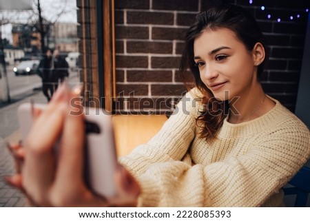 Smiling woman dressed warm sweater, hold phone gadget and taking selfie in street cafe. Lady have a chat with friends, video call meeting, talking speaking