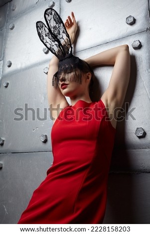 Young woman in black rabbit or hare fancy mask and red dress. Eyes closed. Hands up. In the background there is a metal wall with rivets . Royalty-Free Stock Photo #2228158203