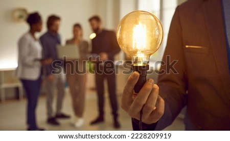 Man in suit holding bright electric Edison light bulb as symbol of developing creative business idea. Banner. Close up of glowing lightbulb in human hand, diverse team of workers in blurred background