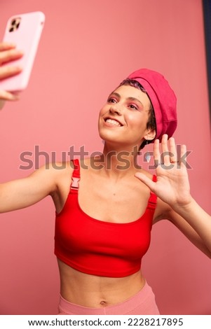 A young woman blogger with colored pink hair and a short haircut takes a picture of herself on the phone and broadcasts a smile in stylish clothes and a hat on a pink background monochrome style