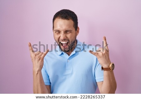 Hispanic man standing over pink background shouting with crazy expression doing rock symbol with hands up. music star. heavy music concept. 