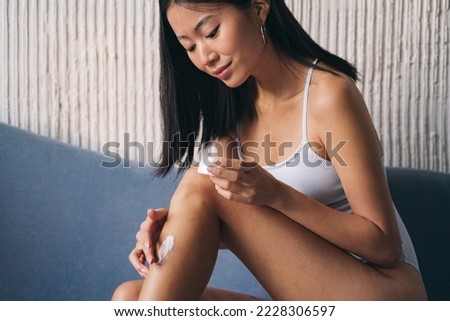 Young asian woman applying cream on her leg as part of body care morning routine, sitting on couch at home
