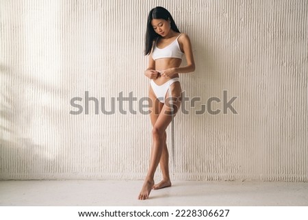 Asian woman measuring waist line with tape measure against textured wall. Korean fitness girl doing body measurement during healthy lifestyle marathon, checking weight loss