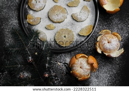 sweet orange juicy tangerines in a peeled peel lie on a black background and a black kitchen tray next to a spruce Christmas branch and sprinkled with white powder.for cards, banners, flyers, invitati