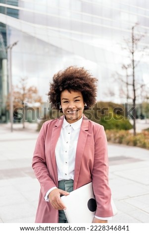 Pretty black business woman with afro hair holding her laptop and looking at camera. She is walking in a business area of the city.