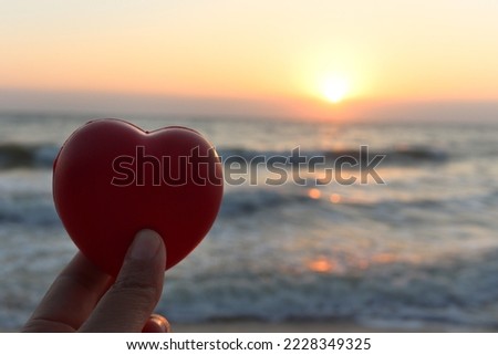 Hand holding red heart with beautiful orange sunset sky on the tropical sea beach, Nature Love Concept