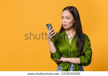 Displeased puzzled young brunette woman of Asian ethnicity wearing basic green satin shirt hold in hand use mobile cell phone read fake news isolated on bright yellow colour background studio portrait