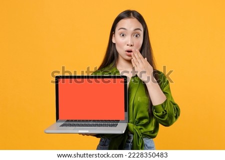 Young IT woman of Asian ethnicity wear basic green satin shirt hold use work on laptop pc computer with blank empty screen mock up copy space cover mouth isolated on yellow background studio portrait