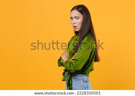 Side view sad upset tired young brunette woman of Asian ethnicity 20s she wear basic green satin shirt stand hold hands crossed look camera isolated on bright yellow colour background, studio portrait Royalty-Free Stock Photo #2228350501