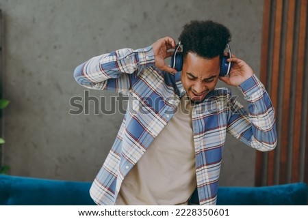 Young cool man 20s wears casual shirt headphones dance near blue sofa listen to music stay at home hotel flat rest relax spend free spare time in living room indoors grey wall People lounge concept
