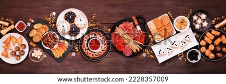New Years Eve theme appetizer table scene. Top view on a dark wood banner background. Charcuterie board, champagne, clock cookies, tuxedo brie cheese and a selection of party food. Royalty-Free Stock Photo #2228357009