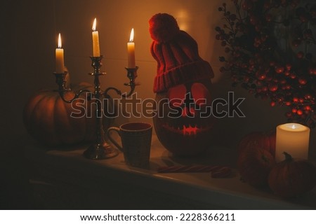 Halloween pumpkin smiling jack lantern and burning candlestick by three candles on fireplace at night home interior. Hallows eve decoration funny glow pumpkin with red berry wreath at dark background