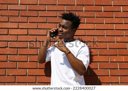 Young black man on the street sitting on the curb with his phone and texting. Portrait of a guy wearing a casual outfit leaning on the red brick wall with copy space for text. Background.