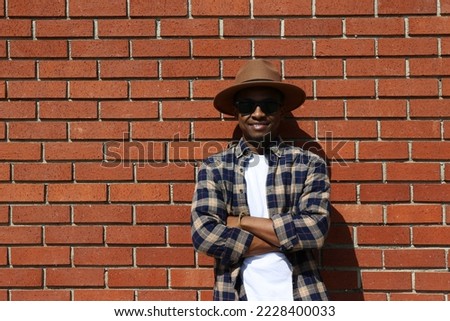 Young black man wearing wide brimmed fedora hat, sunglasses and checked shirt, posing over red brick wall background with copy space. Tanzania born hipster guy leaning on the wall. Royalty-Free Stock Photo #2228400033