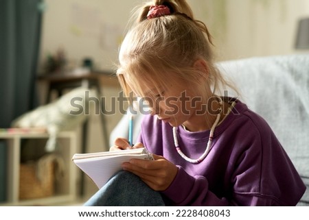 Little girl writing her secrets in her diary while sitting in the bedroom