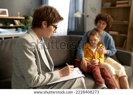 Social worker making notes in document while talking to difficult child and his mom at home Royalty-Free Stock Photo #2228408273