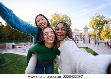 Playful photo of three multicultural female friends taking smiley selfie doing piggyback. Young women having fun together outdoors at city park in Madrid. People enjoying travel in weekend holidays.