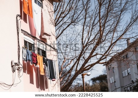 A perspective view of a residential house wall with rows of different clothes drying outside the windows and the bare tree in the defocused background on a bright cold day
