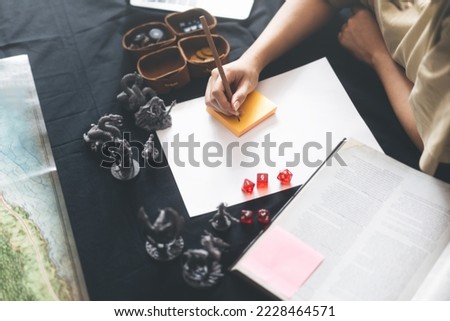 Role playing tabletop and board games hobby concept. Woman hand writing for create story setup adventure sword and magic theme. Foreground with blur monsters miniatures and dice. Royalty-Free Stock Photo #2228464571