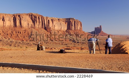 An obese photographer with a wife ready to shade the camera and two children playing nearby is shooting Monument Valley (Navajo Nation) on November morning.