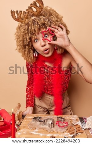 Photo of surprised young woman looks through cutter stares amazed prepares for Christmas holiday bakes gingerbread cookies stands near kitchen table against brown background. Bakery concept.