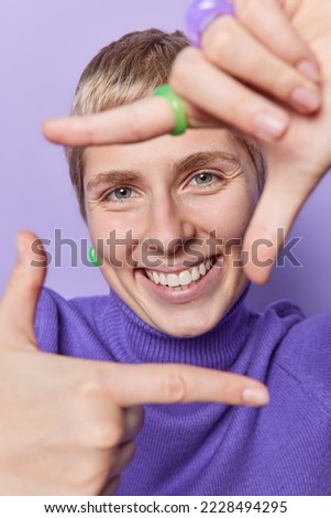 Happy short haired young woman takes photo with frame gesture looks through fingers as if photographing with camera smiles toothily wears purple turtleneck poses indoor searches perspective.