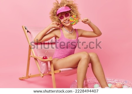 Happy woman sweet tooth poses with lollipop in summer slothes sits on deck chair smiles gladfully enjoys perfect holiday at seaside gets suntan isolated over pink background. Vacation concept