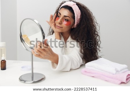 Cute tanned pretty curly Latin lady with pink patch for eye admiring herself doing beauty procedure in white home interior. Copy space. Moisturizing beauty grooming, skincare and pampering concept