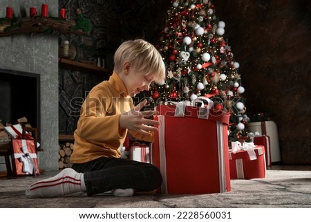 Happy child opens big Christmas gift on Christmas tree background. Joyful boy in cap Santa Claus with Xmas present. New Year time