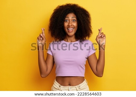 Indoor shot of excited young woman with bushy hairstyle and fingers crossed, makes a wish , smiles toothily, wears purple T-shirt, isolated over yellow background.