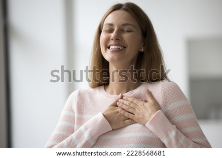 Smiling sincere young 30s woman put folded palms on chest, feeling deeply thankful standing alone indoors. Female showing gratitude, express appreciation, showing symbol of believe and acknowledgement Royalty-Free Stock Photo #2228568681