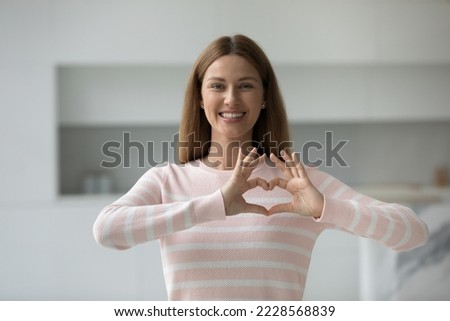 Smiling young woman showing heart sign with fingers, feeling grateful pose alone at home look at camera. Female volunteer expressing support and kindness, make symbol of love, charity and gratitude