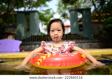 little Asian girl swims in a pool in an orange life preserver.